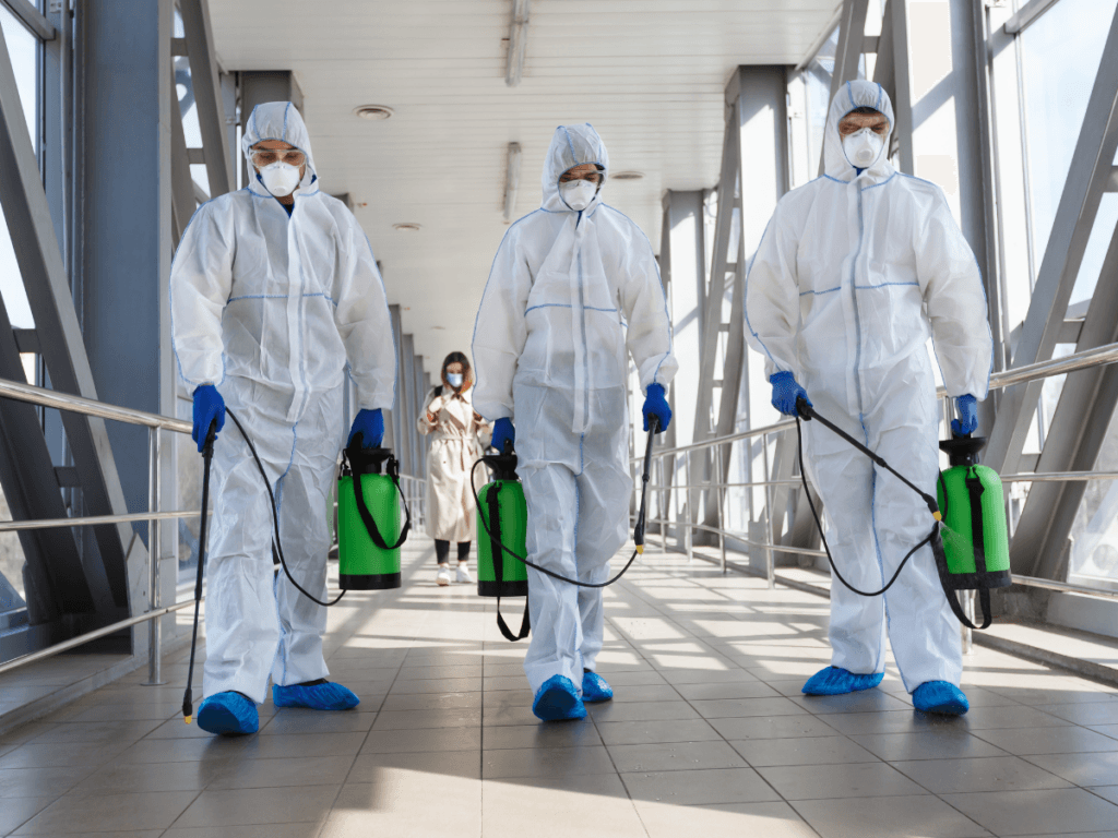 Three people in full protective suits and masks walk indoors holding green disinfectant sprayers, demonstrating the disinfecting sanitizing difference; a fourth person stands in the background.