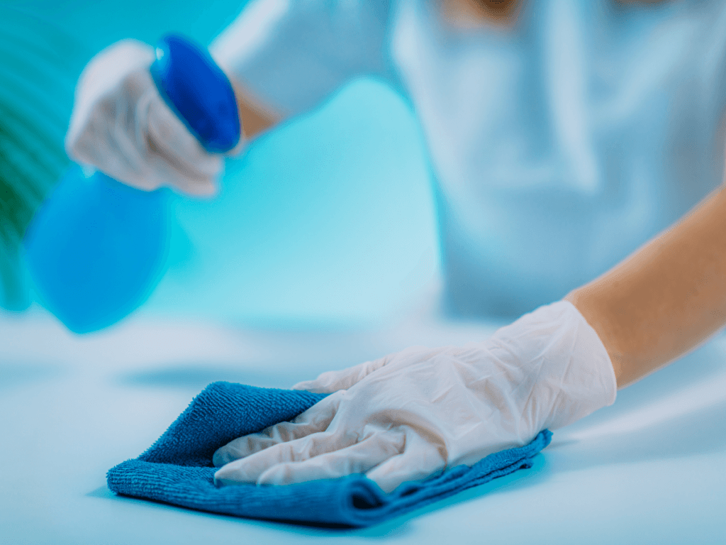 Gloved hands cleaning a surface with a blue cloth and spraying disinfectant, highlighting the disinfecting sanitizing difference.