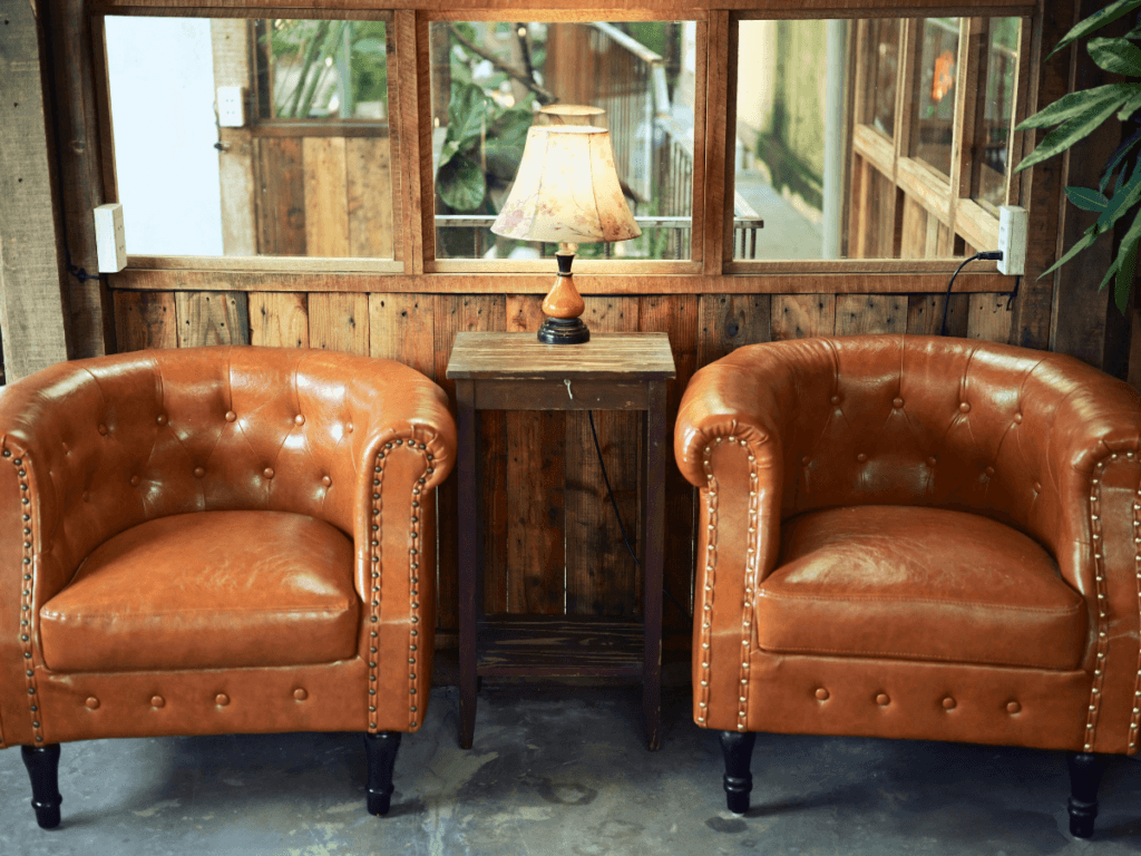 Two brown leather armchairs, part of elegant leather furniture, are placed on either side of a small wooden table with a lamp, set against a wooden wall with windows.