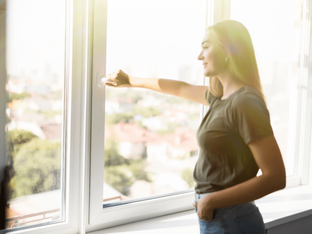 A woman in a t-shirt and jeans stands by a large window, opening it to let sunlight stream in for natural odor removal throughout the room.