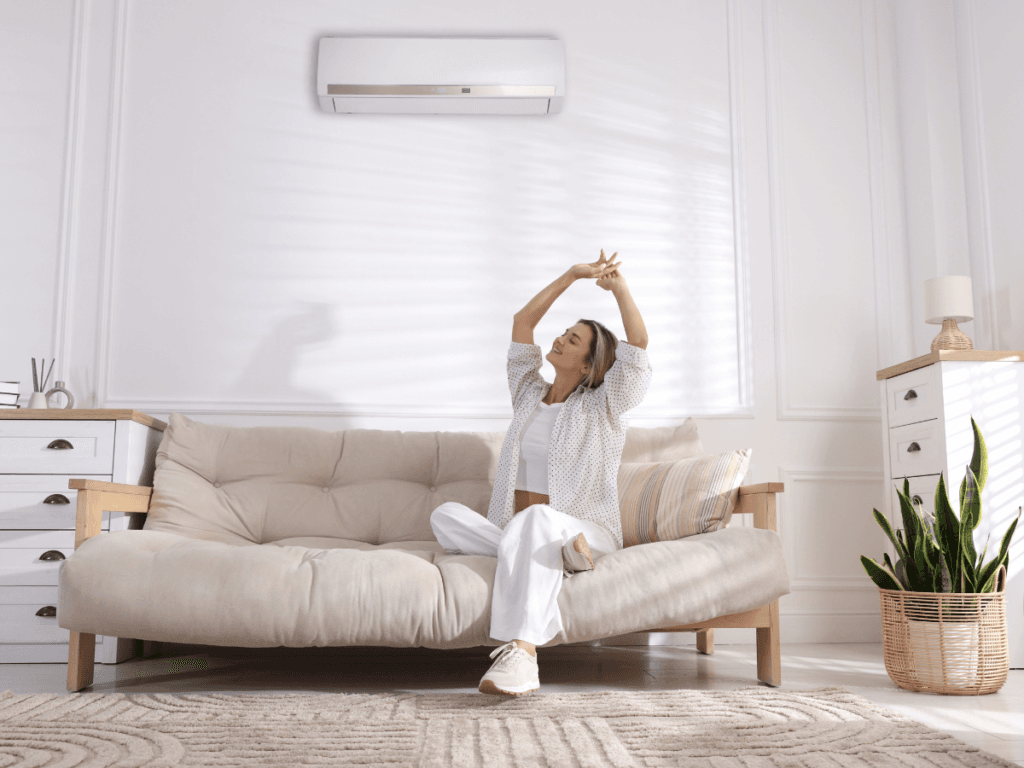 A woman sits on a beige sofa in a bright living room, stretching with her arms raised. An air conditioner with natural odor removal is mounted on the wall above her.