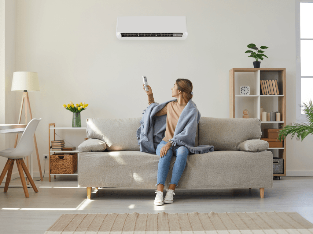A woman sitting on a sofa with a blanket uses a remote to operate an air conditioner with natural odor removal in a bright, modern living room.