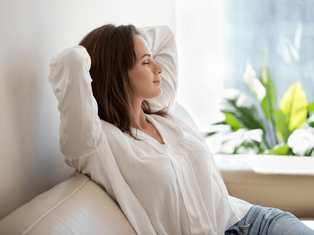 Woman in a white blouse sits on a sofa with eyes closed and arms behind her head, appearing relaxed, enjoying natural odor removal, with a green plant in the background.