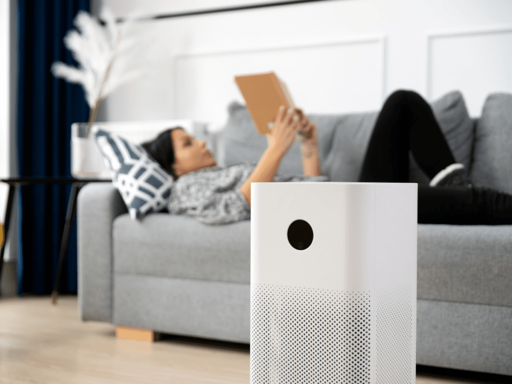 A woman lies on a gray sofa reading a tablet, with a white air purifier known for natural odor removal positioned in the foreground of a modern living room.