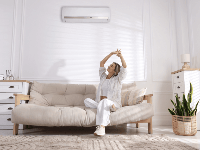A woman sits on a beige sofa in a bright living room, stretching with her arms raised. An air conditioner with natural odor removal is mounted on the wall above her.