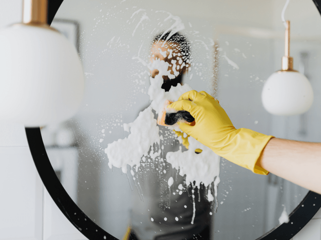 A person wearing a yellow glove speed cleaning a round mirror with a sponge and cleaning foam, their reflection partially visible.
