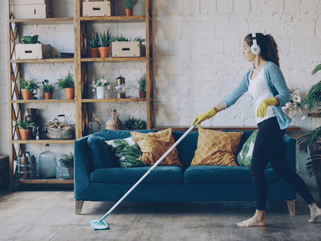 A woman wearing headphones and cleaning gloves speed cleans by mopping the floor in a living room, featuring a blue sofa and a shelf filled with plants and boxes in the background.
