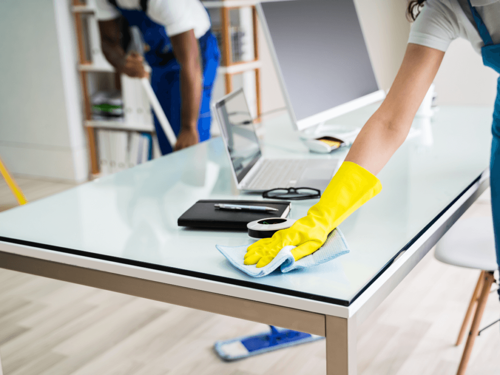Two people clean an office; one wipes a glass desk with a yellow glove, while the other sweeps the floor. Laptops and office supplies are on the desk, showcasing the clean office benefits of improved organization and productivity.