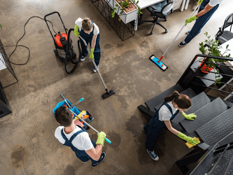 Four people in uniforms clean an office floor with mops and a vacuum, while one person wipes a stair railing; cleaning equipment and plants are visible, highlighting the clean office benefits of a well-maintained workspace.