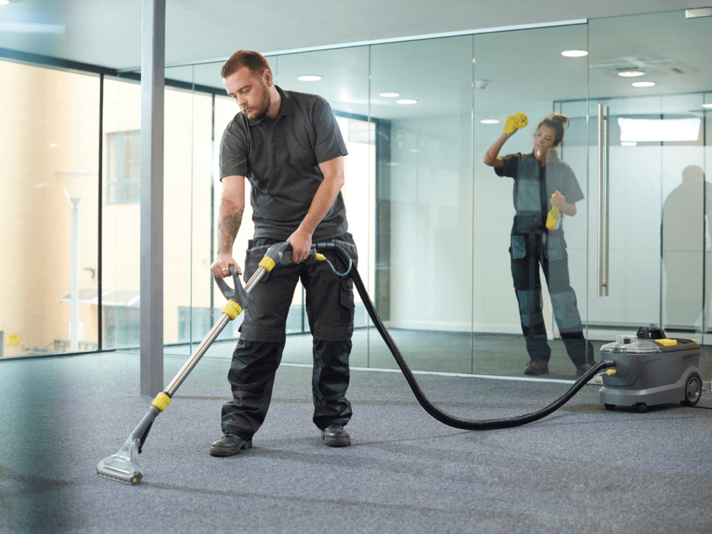 A man vacuums a carpeted floor while a woman cleans a glass wall in an office space with modern furnishings and large windows, highlighting the clean office benefits of maintaining a tidy and organized work environment.