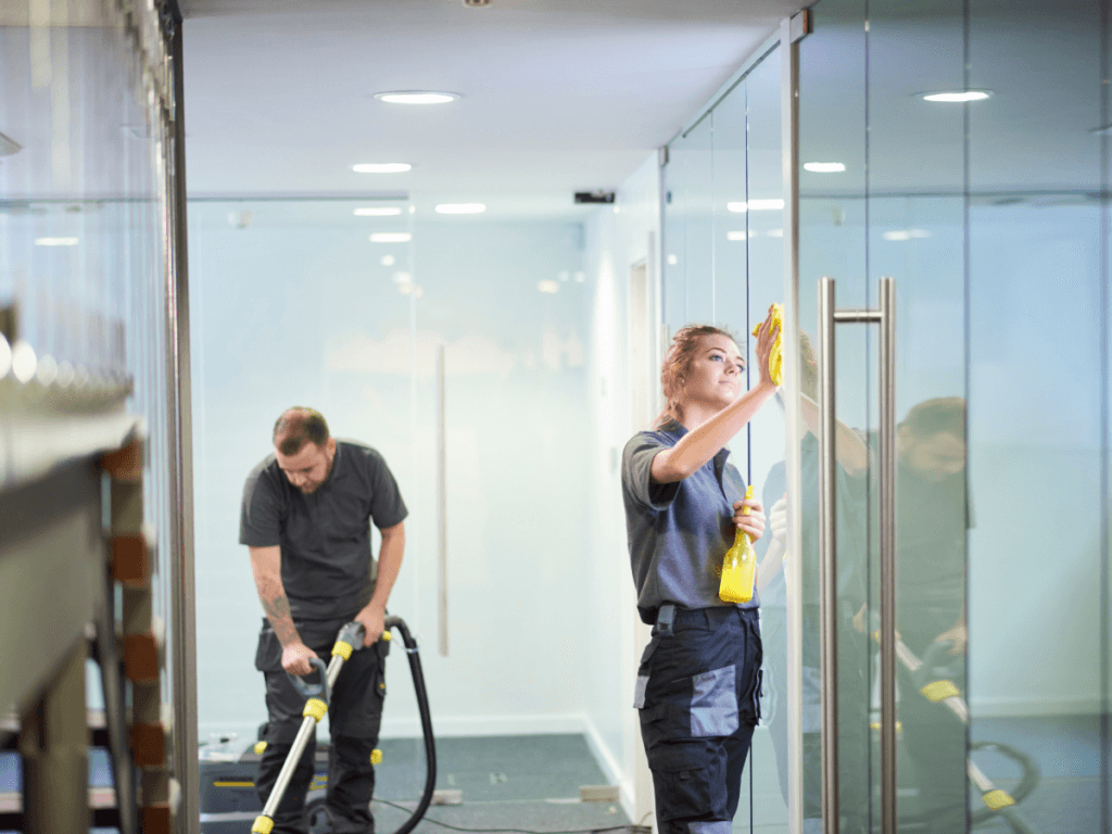 Two people in uniforms clean an office; one vacuums the floor while the other wipes a glass door with a cloth and spray bottle, demonstrating the clean office benefits of a tidy and organized workspace.