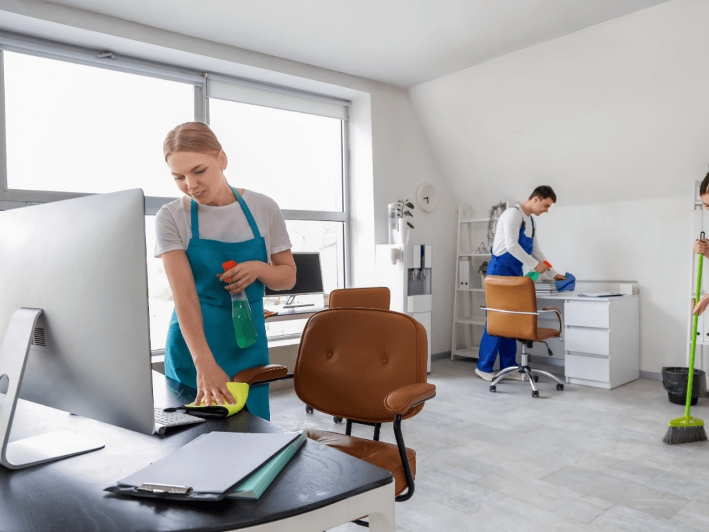 Three people wearing cleaning uniforms are cleaning an office with desks, chairs, and a computer. One wipes a desk, another cleans a table, and the third sweeps the floor&mdash;demonstrating the clean office benefits of improved organization and a healthier work environment.