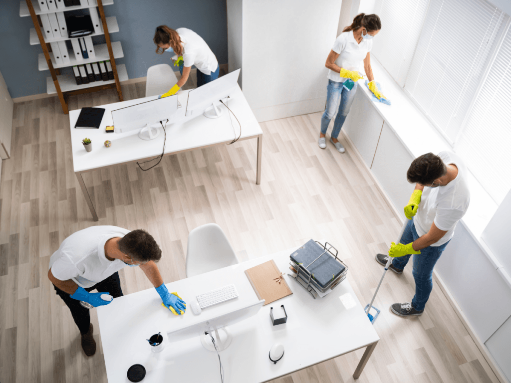 Four people in gloves and white shirts clean an office, wiping desks and windows and mopping the floor, demonstrating the many clean office benefits such as improved productivity and a healthier work environment.
