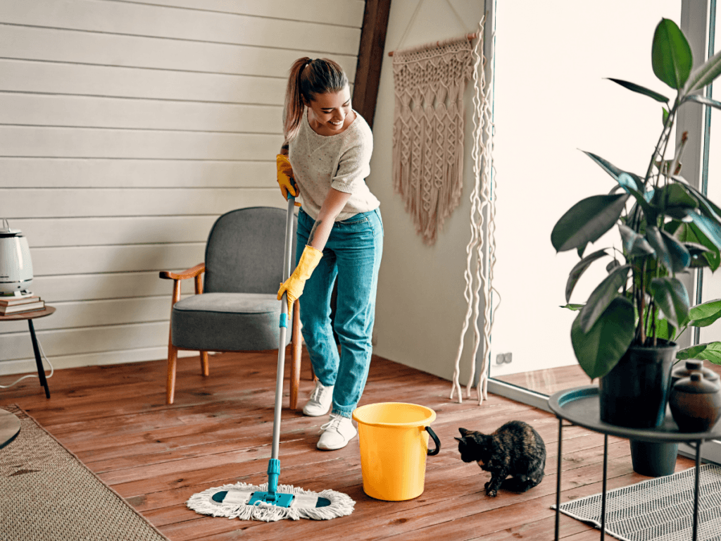 Woman wearing gloves mops a wooden floor in a living room with a yellow bucket nearby; a black cat sits beside her, ensuring the space is pristine and ready for vacation rental cleaning.