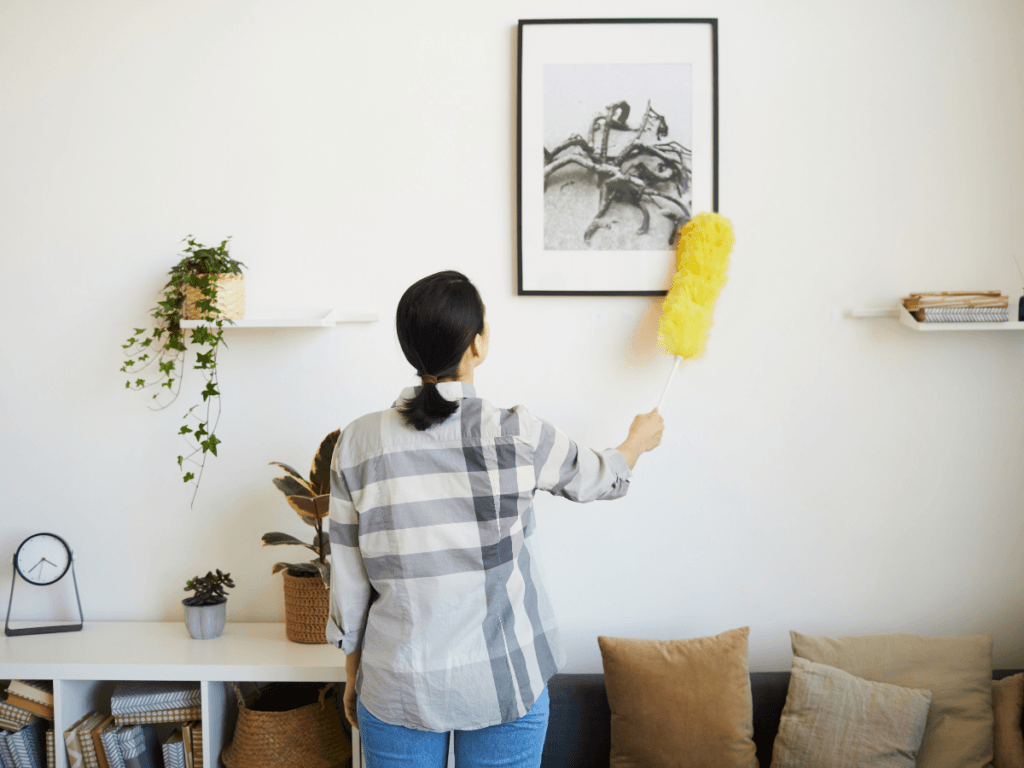 Person using savvy dusting techniques to clean a framed picture on a white wall with a yellow duster; plants, shelves, and a clock decorate the room.