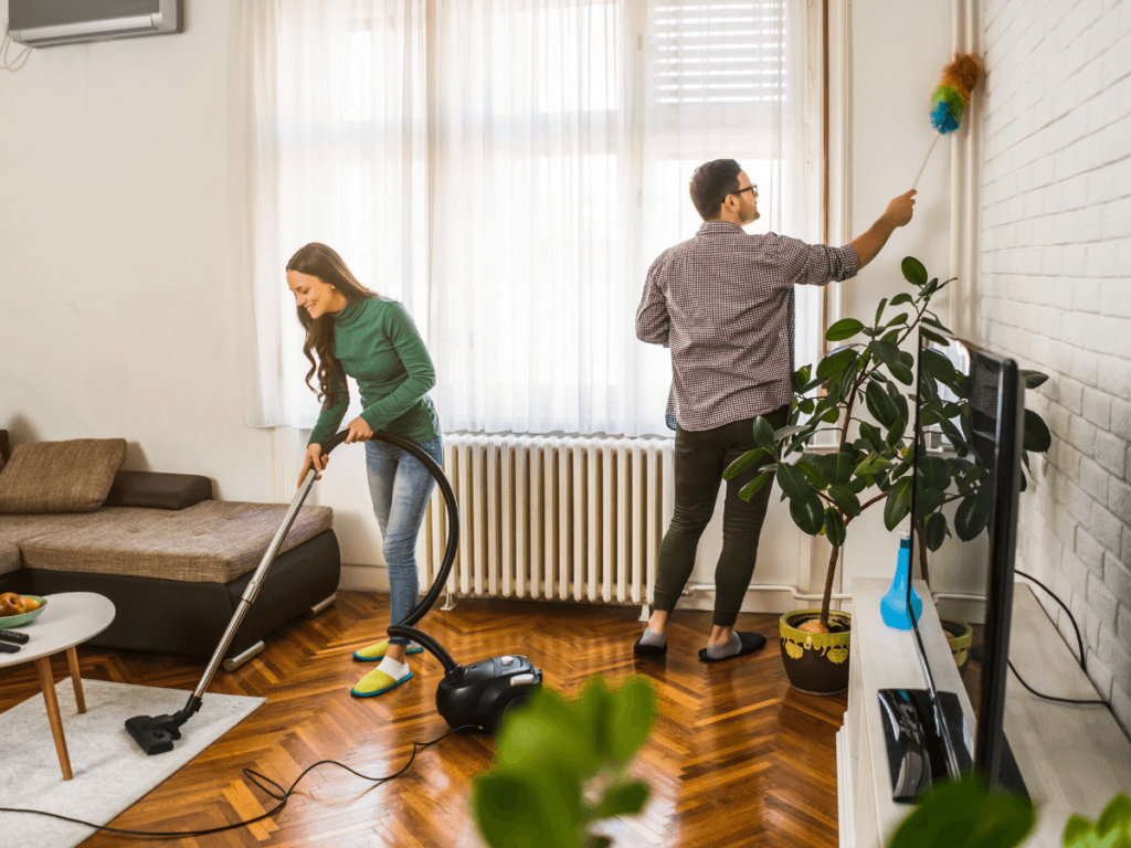 A woman vacuums the living room floor while a man demonstrates effective dusting techniques, using a feather duster to clean the wall.
