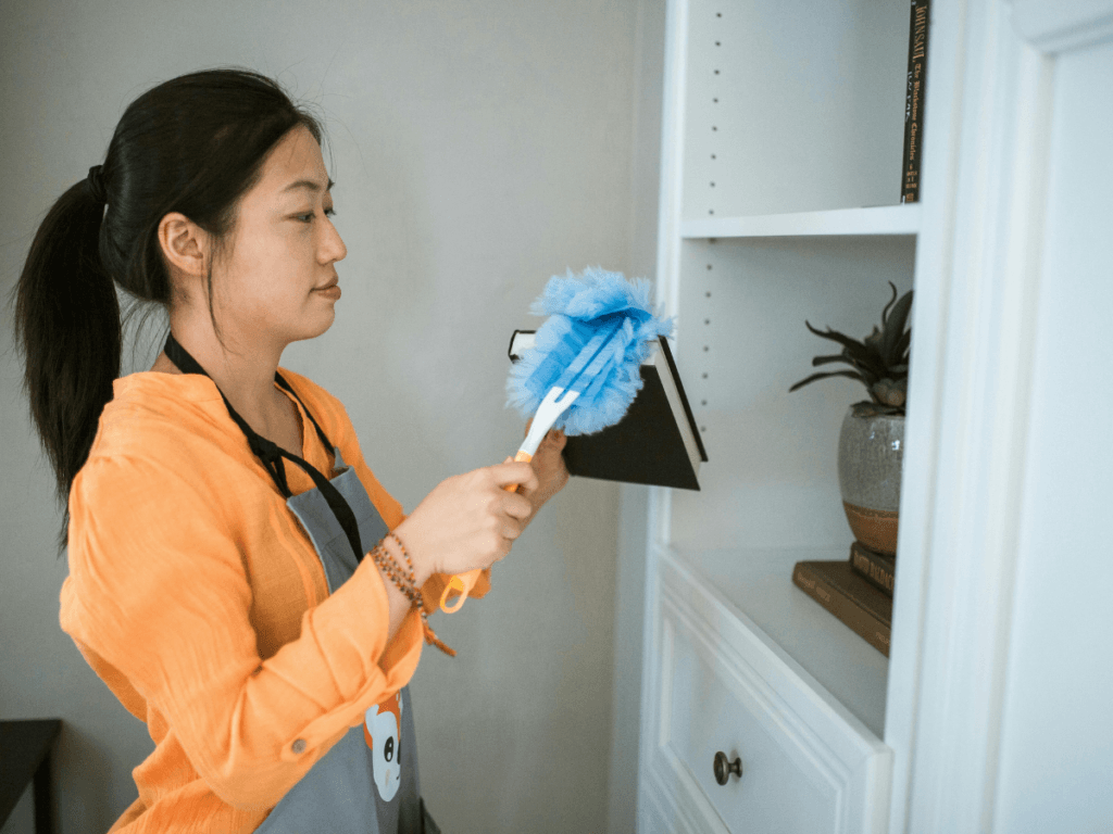 A woman in an orange shirt demonstrates effective dusting techniques as she dusts a white bookshelf with a blue duster. Books, a small plant, and a decorative item are visible on the shelf.
