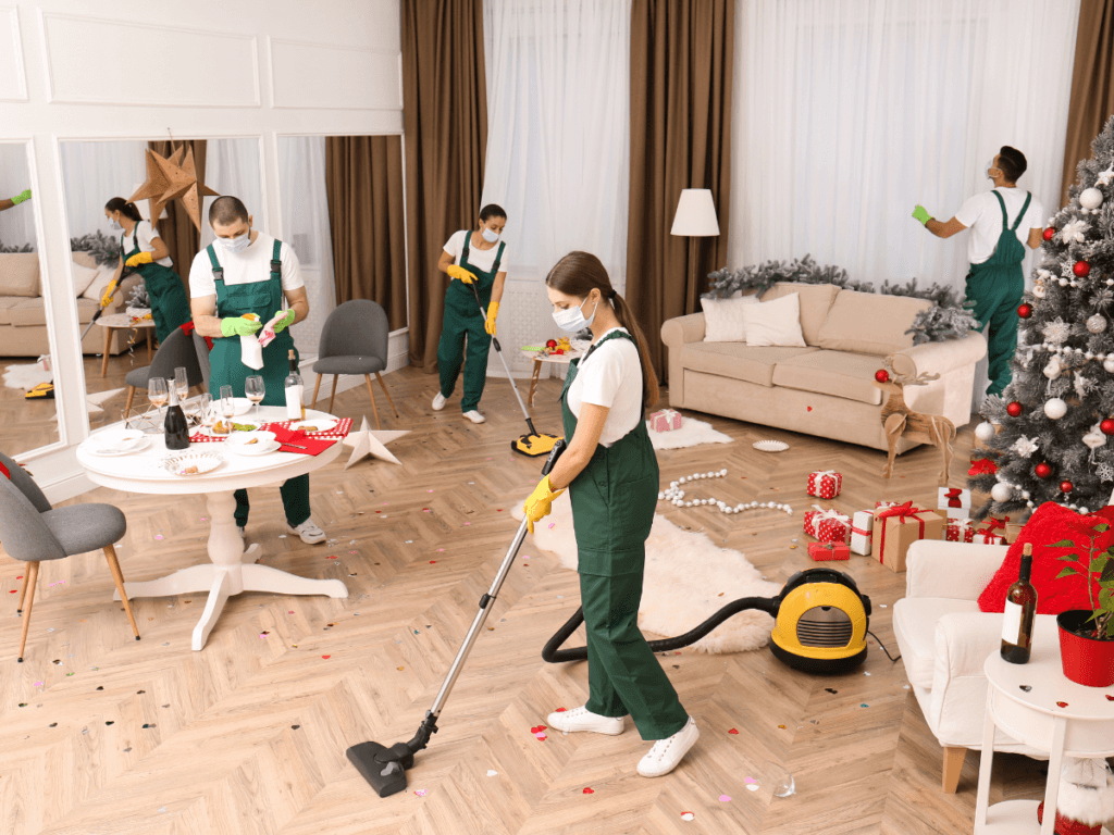 Four people in green uniforms and masks perform event cleanup, tidying a living room with holiday decorations, a Christmas tree, and scattered confetti using vacuum cleaners and cloths.