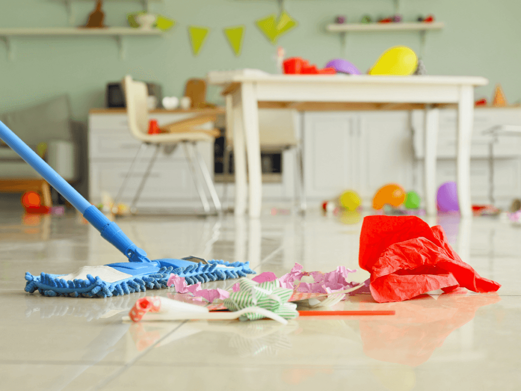 A blue mop and scattered party trash are on a tile floor in a kitchen, evidence of recent event cleanup, with a messy table and balloons in the background.