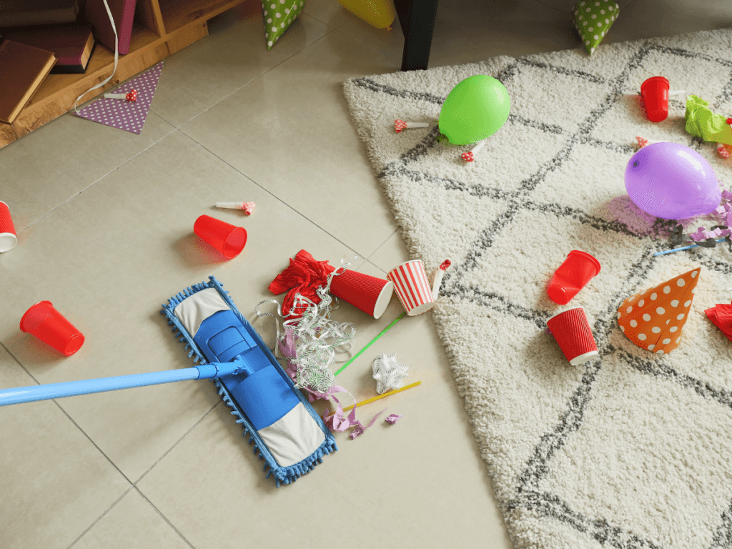 A mop handles event cleanup on a tiled floor scattered with balloons, party hats, red cups, and other party debris next to a carpet.