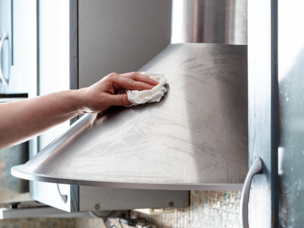 A hand performs stainless steel cleaning on a kitchen range hood, wiping away smudges and streaks with a white cloth.