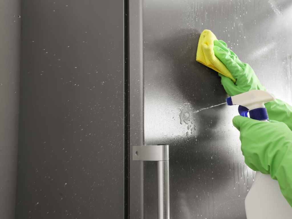 Person wearing green gloves sprays and wipes a glass door with a yellow cloth, cleaning the surface just like during thorough stainless steel cleaning.