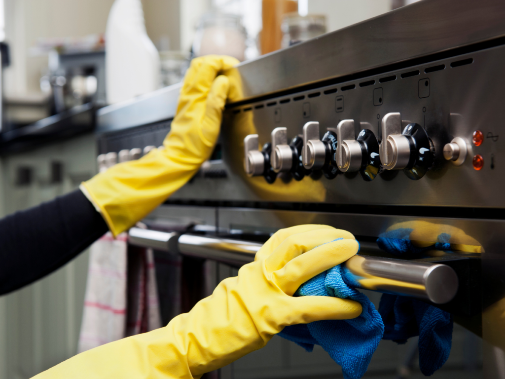 Person wearing yellow gloves performs stainless steel cleaning on an oven with a blue cloth, focusing on the control knobs and handle in a kitchen setting.